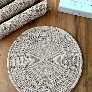 May include: A beige crocheted round placemat with a circular pattern. The placemat is on a wooden surface.