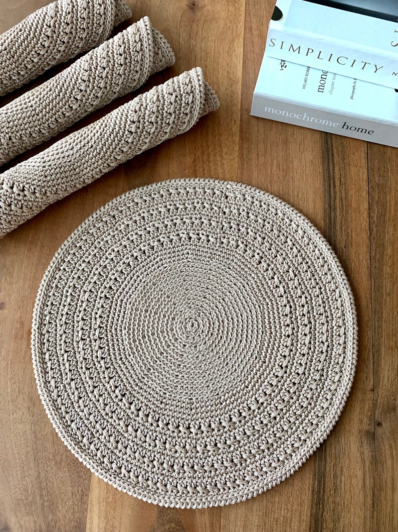 May include: A beige crocheted round placemat with a circular pattern. The placemat is on a wooden surface.