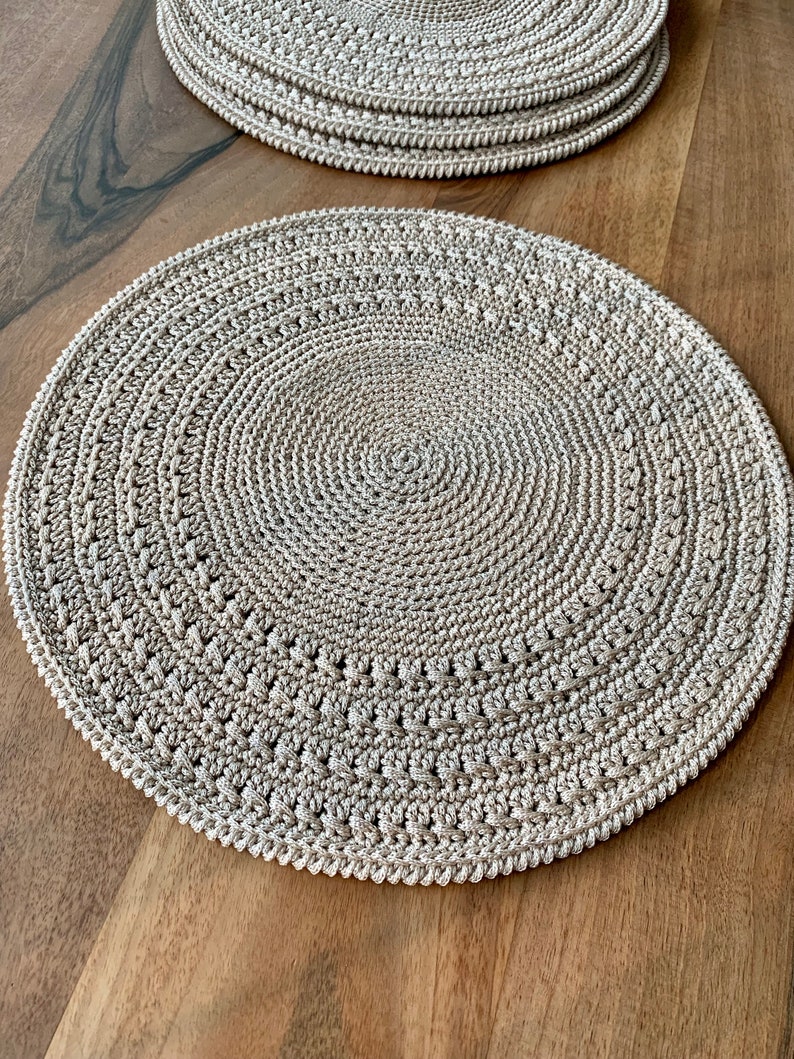 May include: A beige crocheted placemat with a circular design. The placemat is made of a textured yarn and has a slightly raised edge.