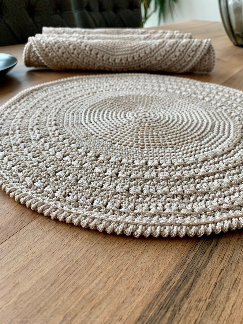 May include: A beige crocheted round placemat with a textured design. The placemat is on a wooden table.