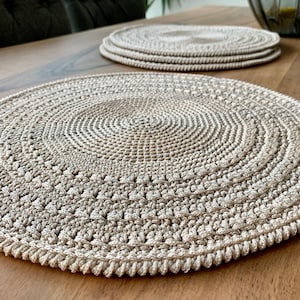 May include: A set of three beige crocheted placemats on a wooden table. The placemats are round and have a textured design.