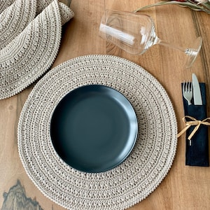May include: A beige crocheted placemat with a black plate on top. A glass wine glass and a black napkin with a fork and knife are on the table.