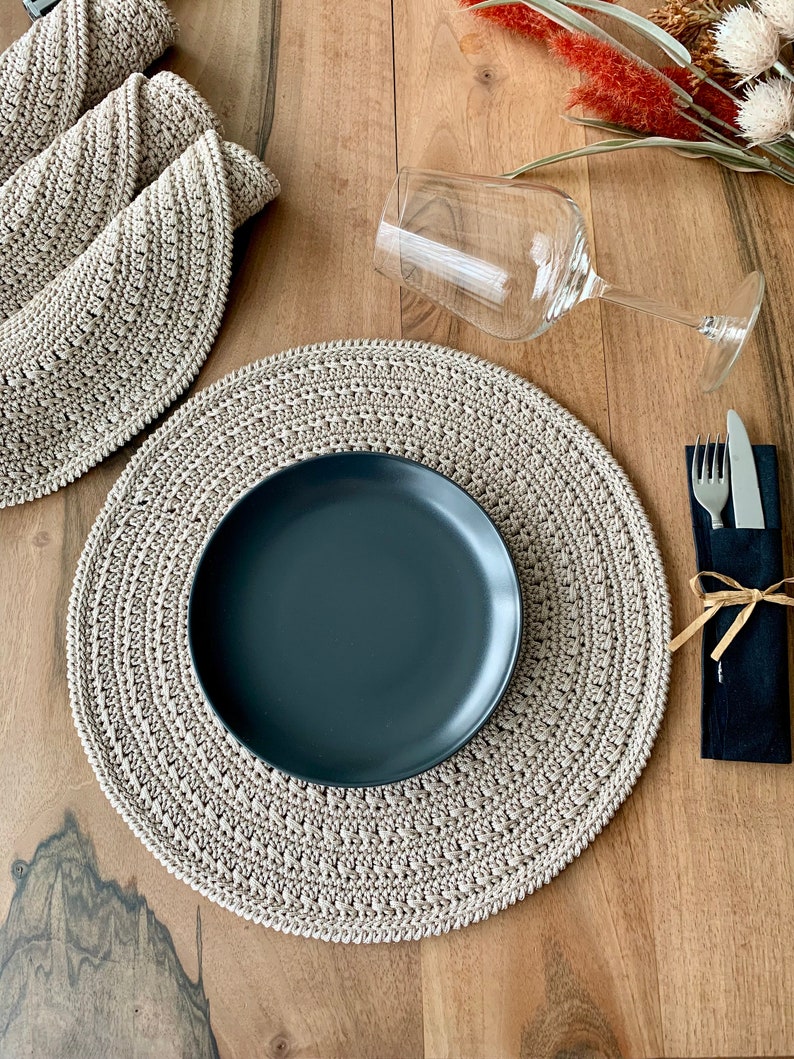 May include: A beige crocheted placemat with a black plate on top. A glass wine glass and a black napkin with a fork and knife are on the table.
