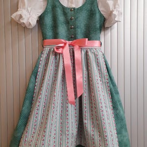 Könnte beinhalten: Ein traditionelles grünes Dirndlkleid mit weißer Bluse und rosa Schleife. Das Kleid hat einen geblümten Rock und ein Mieder mit dekorativen Knöpfen. Die Ärmel sind kurz und gepufft, und das Kleid hängt an einem Holzbügel.