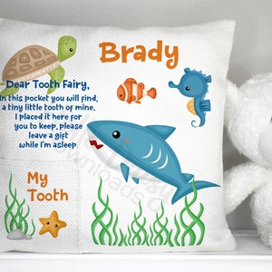 May include: A white pillow with a pocket for a tooth. The pillow has a blue shark, a seahorse, a turtle, and a starfish on it. The text on the pillow reads "Dear Tooth Fairy, In this pocket you will find, a tiny little tooth of mine. I placed it here for you to keep, please leave a gift while I'm asleep. My Tooth Brady".