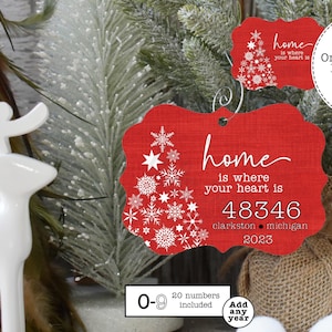 Può includere: Addobbo natalizio rosso e bianco con un motivo a fiocco di neve e il testo "home is where your heart is" con un codice postale, una città e uno stato. L'addobbo include anche l'anno 2023. L'addobbo è su uno sfondo di juta con una statuetta di renna bianca.