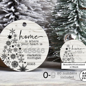 May include: White wooden round ornament with a black snowflake design and the text "home is where your heart is 48346 clarkston michigan 2023".