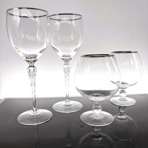 May include: Four clear glass wine glasses with silver rims and decorative stems. Two glasses have tall, twisted stems, while the other two have shorter, rounded bowls. The glasses are arranged on a dark surface.