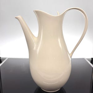 May include: A smooth, off-white ceramic pitcher with a tall, slender neck, a wide rounded base, a curved spout, and a long, thin handle. The surface has a glossy finish that reflects light, creating a clean, uniform appearance throughout the vessel.