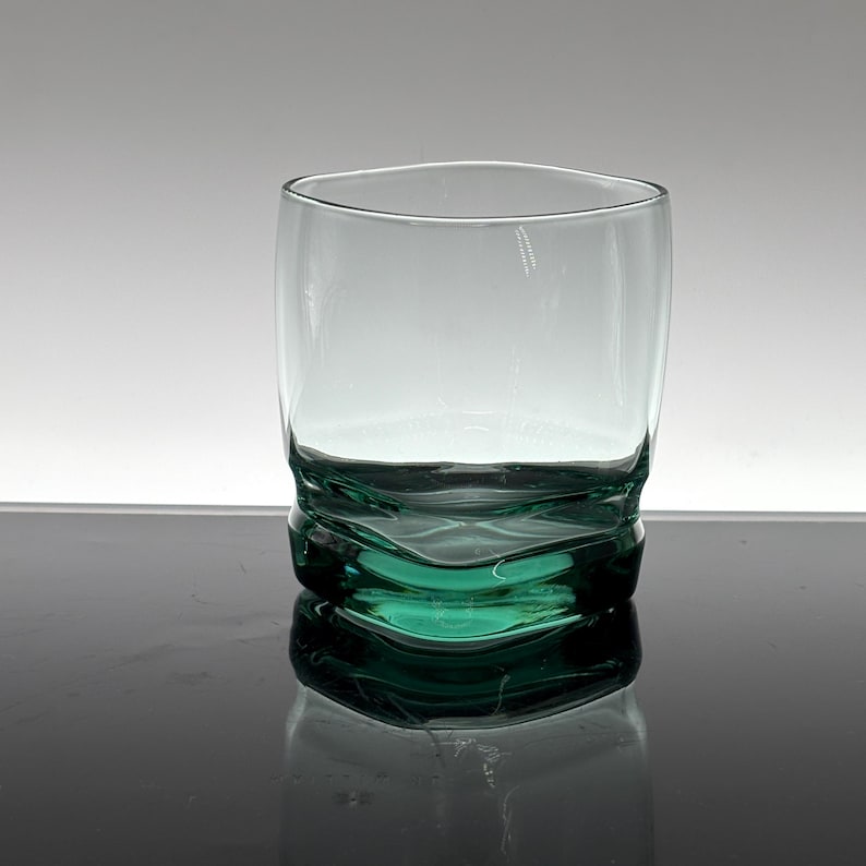 Pu&ograve; includere: Un bicchiere trasparente soffiato a mano con una base strutturata unica di colore verde acqua intenso. Il bicchiere ha una forma arrotondata e una parte superiore liscia e trasparente. La base presenta un design ondulato e organico. Adatto alle bevande.