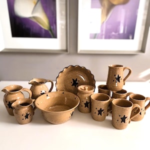May include: A collection of speckled tan ceramic tableware, including pitchers, bowls, plates, mugs, and a lidded jar. Each piece is adorned with a black star design. The set is arranged on a white surface.