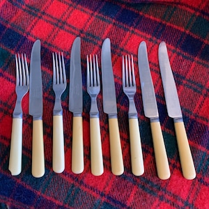 May include: Eight silver forks with ivory handles. The forks are arranged in a row on a red and black plaid surface.