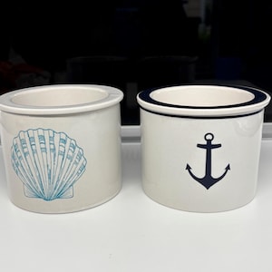 May include: Two ceramic containers with a nautical theme. One features a blue seashell design, and the other has a navy blue anchor. Both have a white base and a light blue rim. The containers are cylindrical.