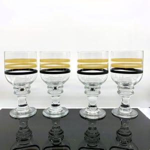 May include: Four clear glass wine glasses with black and gold bands around the top. The glasses have a wide bowl and a tall stem.