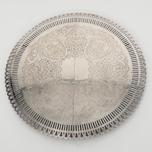 May include: A silver-colored, round serving tray with intricate floral and scrollwork patterns. The tray has a scalloped edge with a pierced design. The tray is divided into two hinged sections.
