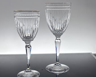 May include: Two clear crystal wine glasses with vertical ribbed detailing and gold rims. The stems have a faceted design, and the glasses sit on round bases. The glasses are elegant and suitable for formal occasions.