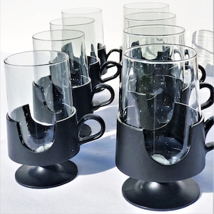 May include: A set of eight clear glass cups with black holders and handles. The cups are cylindrical and sit in a black base with a handle. The holders have a pedestal base. The cups are arranged on a white surface.