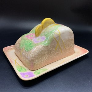 May include: A vintage ceramic butter dish with a floral design. The dish has a cracked glaze finish and features a lid with a yellow flower design. The dish is decorated with pink, purple, and green flowers.