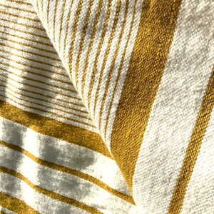 May include: Close-up of a white and mustard yellow striped fabric. The fabric is woven with a textured pattern and has a soft, cozy feel.