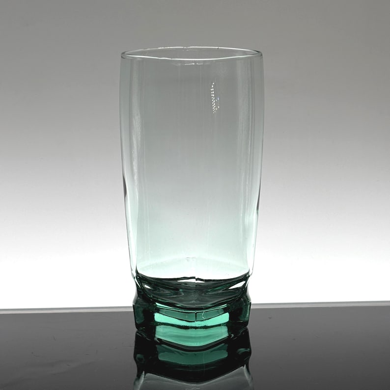 Pu&ograve; includere: Un bicchiere alto e trasparente con una leggera sfumatura verde. Il bicchiere ha una forma cilindrica e una base geometrica. Il bicchiere vuoto sembra essere fatto di un materiale spesso e resistente.