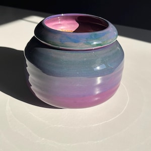 May include: A ceramic jar with a lid, featuring a gradient of colors from purple to blue. The lid is a darker shade of blue and purple, with a glossy finish. The jar is round and has a smooth surface.