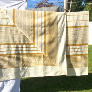 May include: A white and yellow striped throw blanket with a woven texture. The blanket is folded in half and hanging on a clothesline.