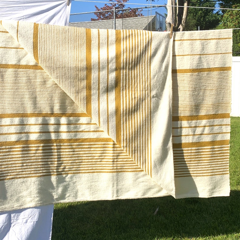 May include: A white and yellow striped throw blanket with a woven texture. The blanket is folded in half and hanging on a clothesline.