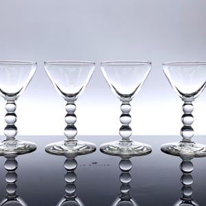 May include: Four clear glass cocktail glasses with a unique design. Each glass features a round bowl, a stem composed of stacked glass spheres, and a decorative base. The glasses are arranged in a row, reflecting on a glossy surface.