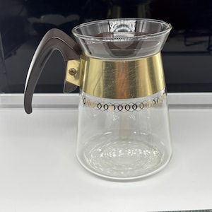 May include: A vintage glass coffee carafe with a brown handle and lid. The carafe has a gold-colored band around the middle with a decorative gold pattern. The carafe is clear, allowing the contents to be visible.