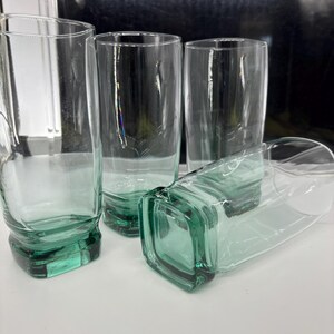 Pu&ograve; includere: Quattro bicchieri trasparenti con base quadrata e una leggera sfumatura verde. I bicchieri sono alti e cilindrici, con un design semplice e moderno. Un bicchiere &egrave; sdraiato su un lato.