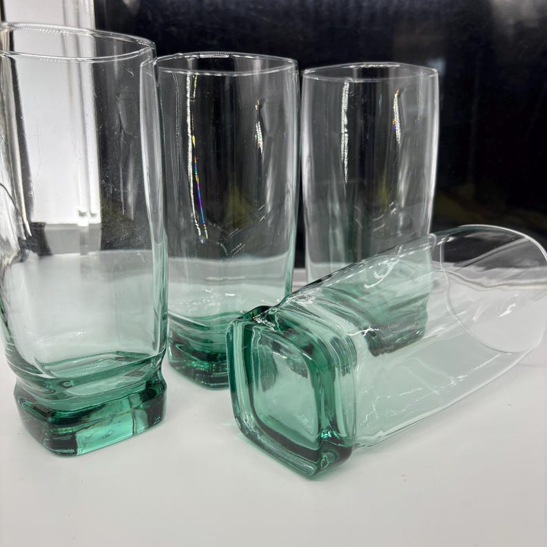 Pu&ograve; includere: Quattro bicchieri trasparenti con base quadrata e una leggera sfumatura verde. I bicchieri sono alti e cilindrici, con un design semplice e moderno. Un bicchiere &egrave; sdraiato su un lato.