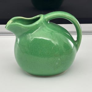 May include: A small, round, green ceramic pitcher with a handle and a spout. The pitcher has a crackle glaze finish. The handle is arched and attached to the body of the pitcher. The pitcher is likely used for serving liquids.
