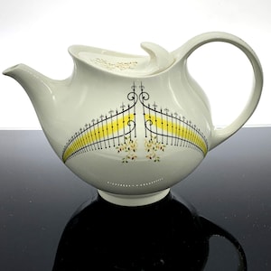 May include: Cream-colored ceramic teapot with a black and yellow gate design. The teapot has a curved spout, a large handle, and a lid with a decorative finial. Floral accents are also present on the teapot.