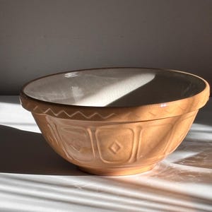 May include: A round ceramic mixing bowl with a light tan exterior and a cream-colored interior. The exterior features a repeating embossed pattern of squares with central diamonds and a zigzag border along the rim. The glaze has a subtle crackle finish.