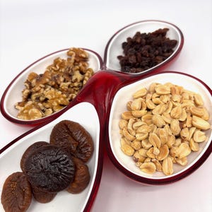 May include: A red and white serving dish with four sections, each filled with different snacks. The snacks include walnuts, raisins, peanuts, and figs. The dish has a unique, clover-like shape.
