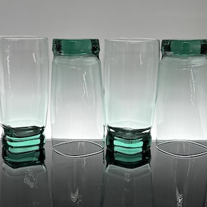 Pu&ograve; includere: Quattro bicchieri trasparenti con base e bordo verde acqua. I bicchieri sono cilindrici con una parte superiore leggermente svasata. La base ha un design impilato e il bordo &egrave; una fascia di colore pieno. I bicchieri sono disposti in fila.