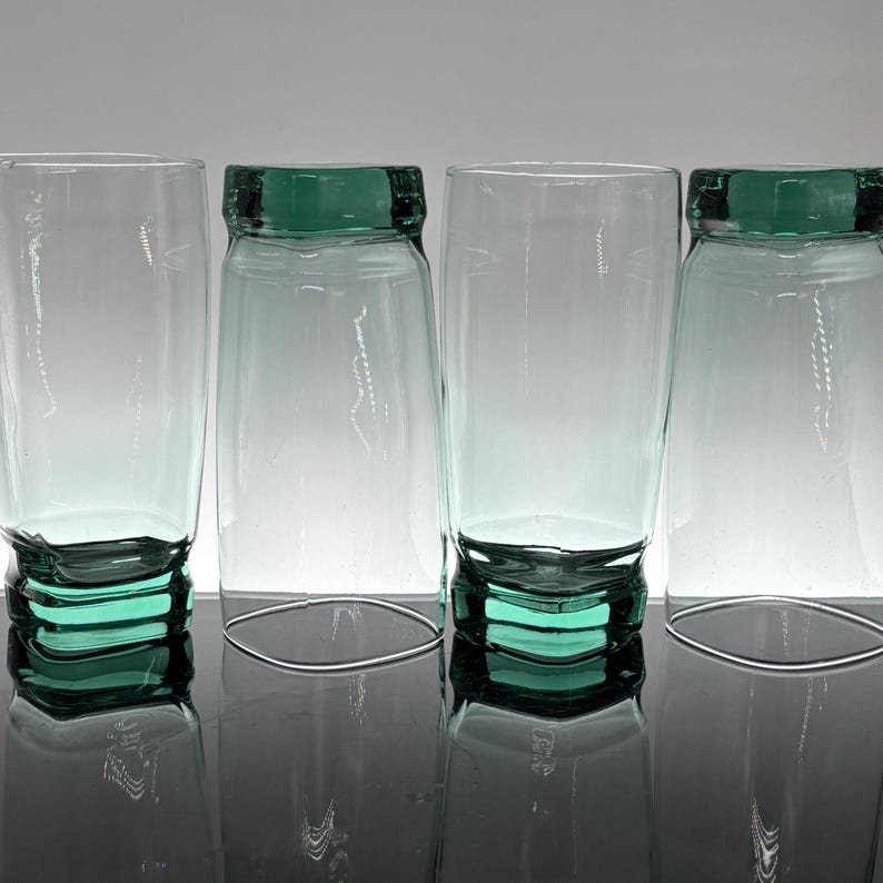 Pu&ograve; includere: Quattro bicchieri trasparenti con base e bordo verde acqua. I bicchieri sono cilindrici con una parte superiore leggermente svasata. La base ha un design impilato e il bordo &egrave; una fascia di colore pieno. I bicchieri sono disposti in fila.