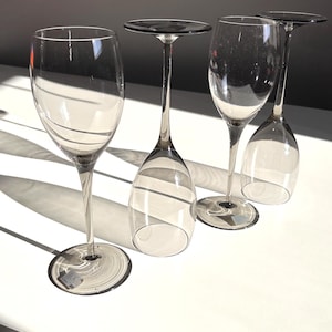May include: Four clear wine glasses with long stems and circular bases are arranged on a white surface. The glasses are partially transparent, allowing light to pass through. One glass is upright, while the others are inverted.