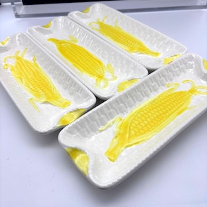 May include: Set of five white ceramic serving trays, each featuring a vibrant yellow corn design. The trays have a rectangular shape with slightly curved edges and a textured basketweave pattern. A bright and cheerful addition to any kitchen.