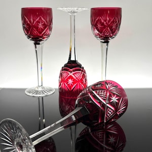 May include: Three wine glasses made of cranberry red cut-to-clear crystal glass. Two glasses stand upright, one is inverted, and one lies horizontally. Each glass features a detailed diamond and starburst pattern on the bowl and a clear stem.