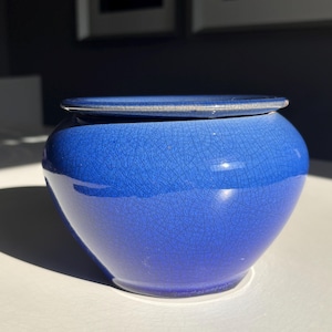 May include: A blue ceramic jar with a lid. The jar has a rounded shape and a crackle glaze finish. The lid is flat and sits on top of the jar. The jar is sitting on a white surface.