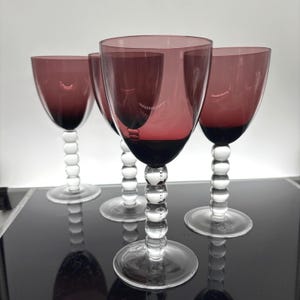 May include: Four wine glasses with a deep red bowl and a clear, bubble-style stem. The glasses sit on a clear, circular base. The wine glasses are arranged in a row, with the light reflecting off the glass.