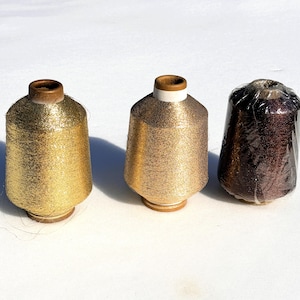 May include: Three spools of metallic thread in gold and brown. The two gold spools are unwrapped, showing the shiny thread wound around a wooden core. The third spool is wrapped in clear plastic and is a darker brown color.