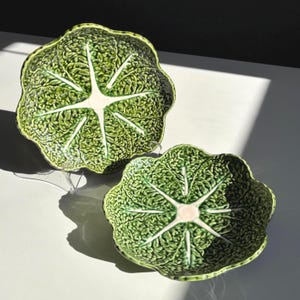 May include: Two green ceramic cabbage-shaped bowls. The bowls have a textured surface with a central white starburst design. The bowls are on a white surface, with shadows cast by sunlight.