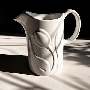May include: A white ceramic pitcher featuring a raised, embossed tulip flower design on its exterior surface. The pitcher has a curved handle on the right side and a distinct pouring spout on the left, with a smooth, glossy finish throughout.