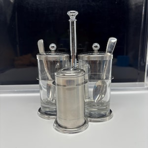 May include: A silver-toned condiment set with three clear glass containers and a central shaker. Each container has a lid and a small spoon. The set is on a silver-toned base and has a central handle.