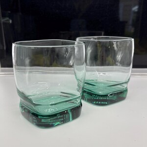 Pu&ograve; includere: Due bicchieri trasparenti di forma quadrata con una base color verde acqua. I bicchieri hanno un design unico e strutturato sul fondo e i lati sono lisci. I bicchieri sono vuoti e sembrano essere di vetro.