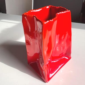 May include: A bright red ceramic vase shaped like a crumpled paper bag. The vessel features a glossy, reflective finish with irregular, indented sides and a serrated, scalloped top edge that mimics the appearance of a folded paper grocery bag.