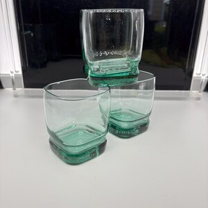 Pu&ograve; includere: Tre bicchieri trasparenti con base quadrata e parte superiore arrotondata. La base di ogni bicchiere ha una tinta verde. I bicchieri sono impilati, due in basso e uno sopra.