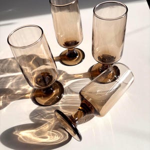 May include: Four amber-colored stemmed glasses with a cylindrical shape. The glasses are arranged on a white surface, casting shadows. The stems and bases are a darker shade of amber.